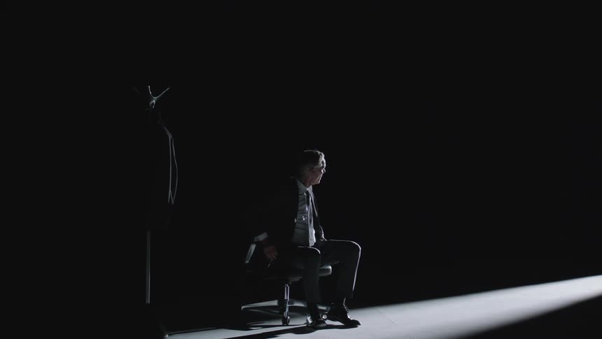 A man sits on a desk chair in the dark illuminated by a beam of light he gets up