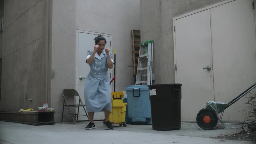 A woman wearing a work uniform dress is standing in next to a building near a mop bucket and ladder with her hands up practicing boxing moves