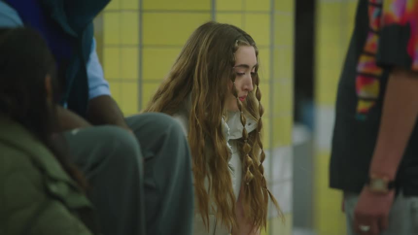Girl with brown hair light blue shirt and ripped jeans sits on bench next to her friends taking off headphones in the subway