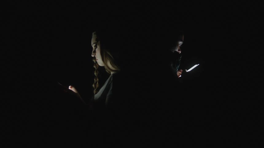 A woman and a man are standing back to back in the dark texting on their cell phones