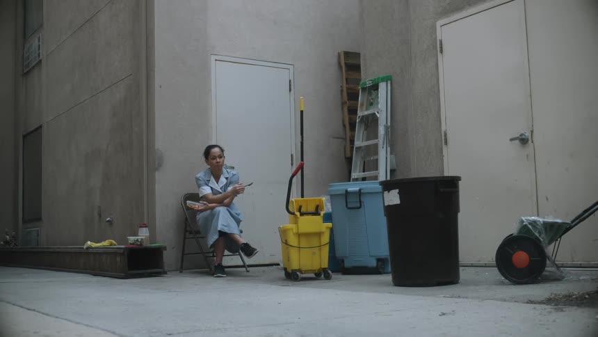 A cleaning woman in uniform sitting outside eating lunch from a container during a break