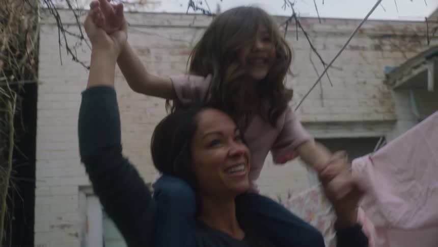 A mother holds her daughter on her shoulders outdoors under a tree near laundry on the clothes lines