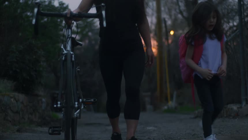 A woman pushing a bicycle talks with a girl as they walk together along a dilapidated road at sunset