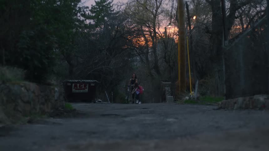 A mother and her daughter walking down an alley while the girl runs around and the woman walks a bike next to her