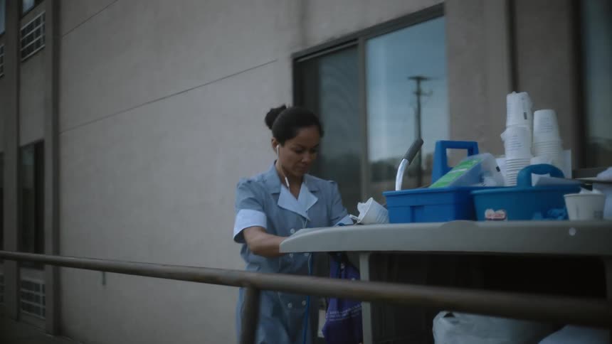 A woman wearing a work uniform dress is walking outside of a building pushing a cart with cleaning supplies