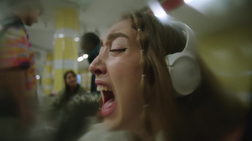A girl wearing headphones is sitting on a train crying and hollering and people are standing in the distance going about their business