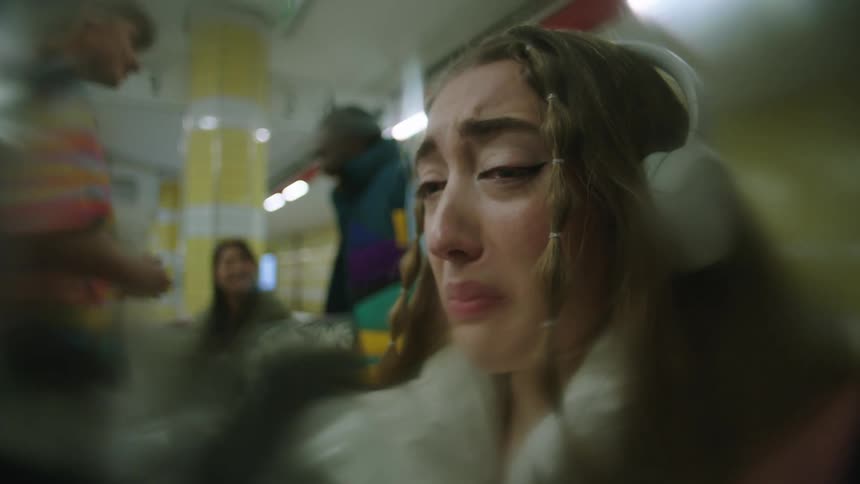 A girl wearing headphones is sitting on a train crying and sobbing and people are standing in the distance talking