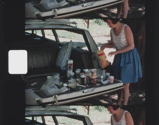 A woman picking up food off the tailgate of a car as she folds a bag of chips