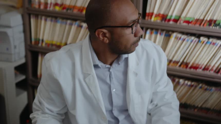 A man in his office surrounded by numerous books he picks up a file and drops it on the table beside him