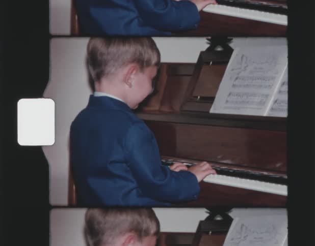 A young boy is playing piano and smiling at the camera positioned behind him