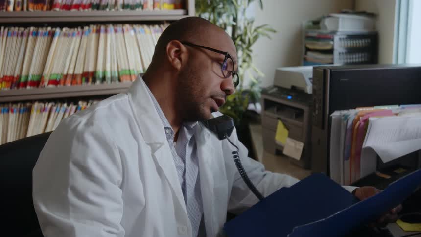 A man looking at a book inside a blue file then dropping it he is engaged in a conversation on the phone he then picks up a yellow file to check its contents