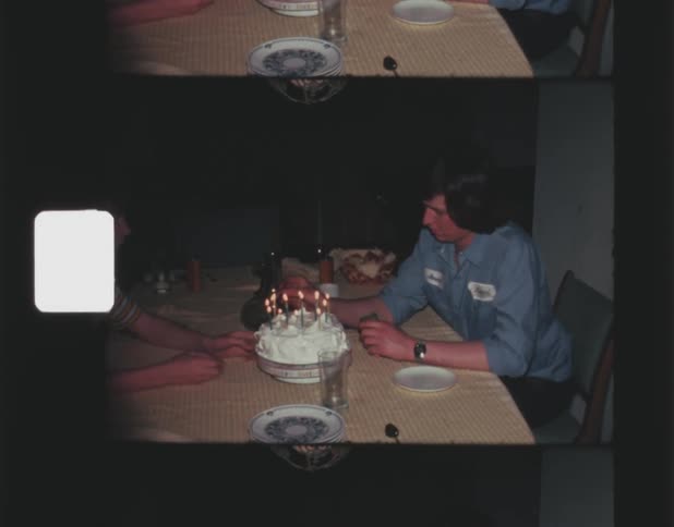 A father and son sit together at a small table with a cake with candles atop it