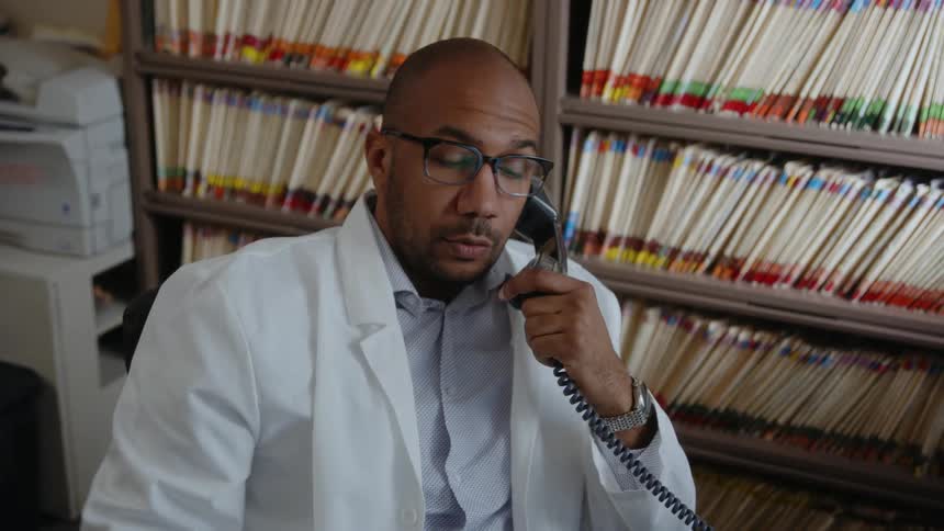 A man wearing a white lab coat is sitting at a desk as he holds a telephone receiver and moves in his chair as he talks