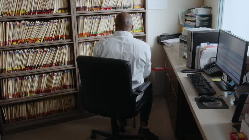 A researcher using a computer in an office stops to retrieve a file from a nearby shelf