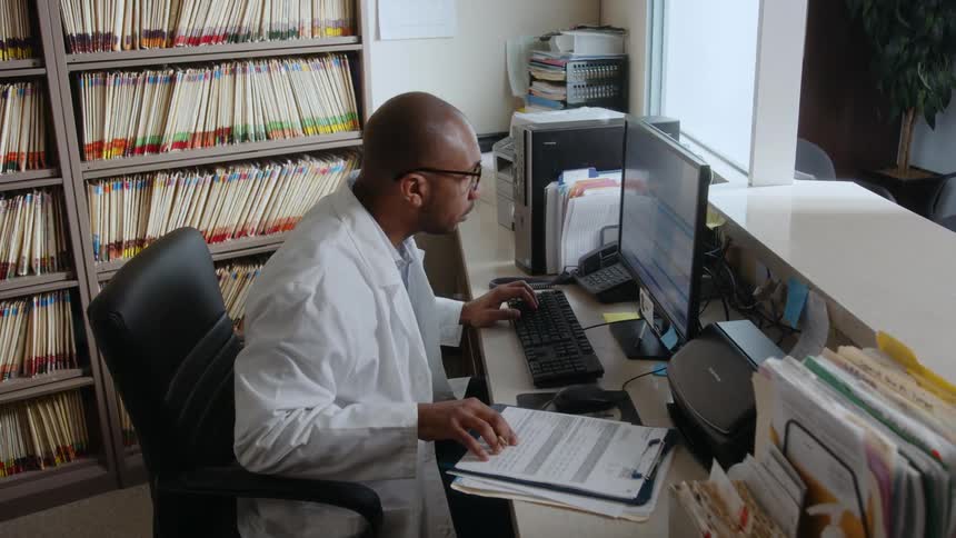 A man wearing a white lab coat is sitting at a desk typing on a computer keyboard and looking at a document as he works