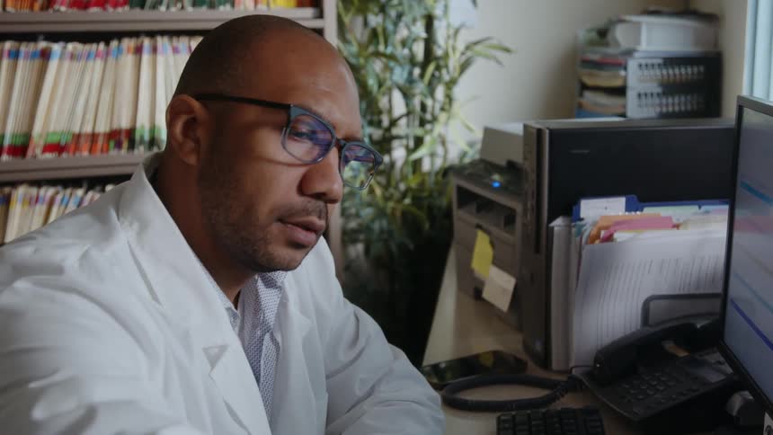A doctor sitting in his office in front of a computer looking at a clipboard