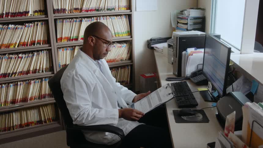 Male doctor sitting in his office chair typing on the keyboard looking at a clipboard then putting it back
