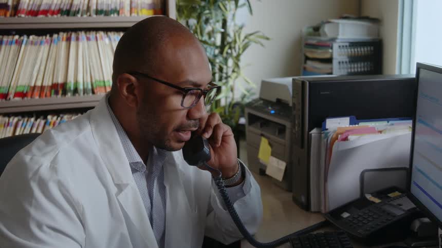 A man wearing a white lab coat is sitting at a desk in front of a computer holding a document as he holds a telephone receiver and talks on the phone