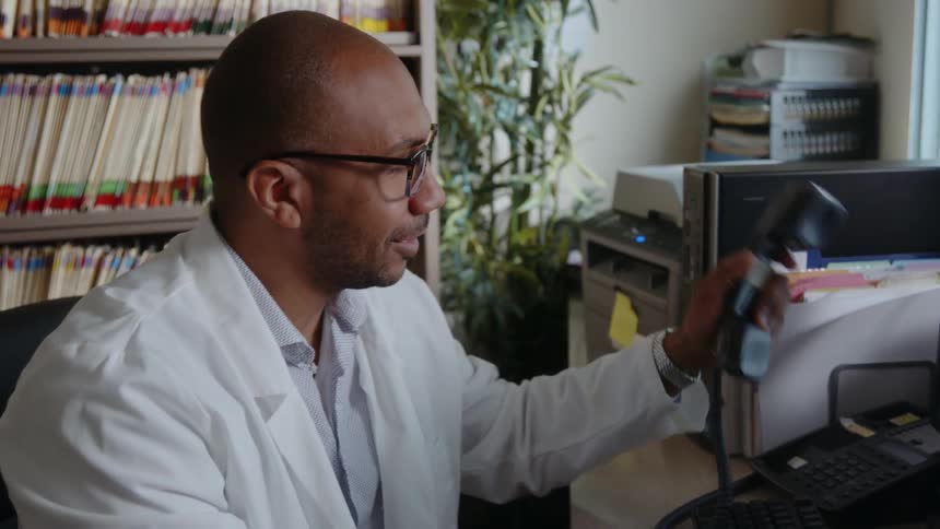 A man wearing a white lab coat smiles as he talks on the phone and hangs up the receiver then looks at papers on his desk