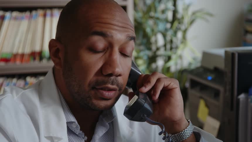 A doctor talks on the phone and takes his glasses off when he gets sad news