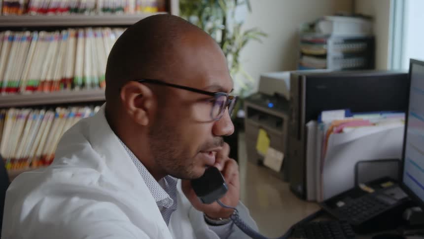 A man in his office engaged in a phone call then grabbing a book to check its contents