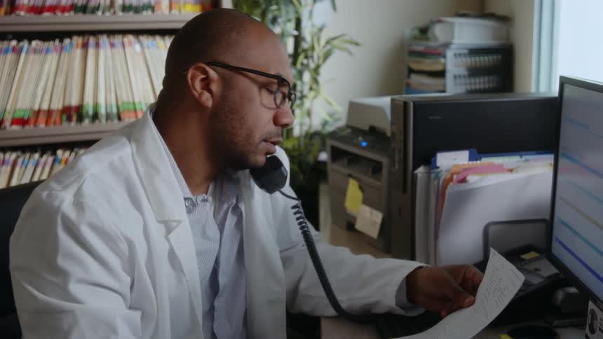 A male doctor sitting in his office holding a paper and dialing the telephone and holding it to his ear