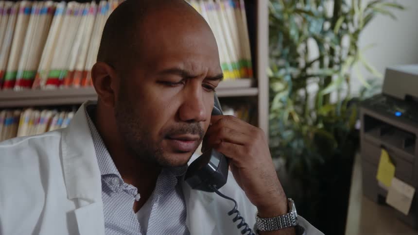 A close up ofa male doctor sitting in his office talking on the telephone then putting his glasses on while sitting up in the chair