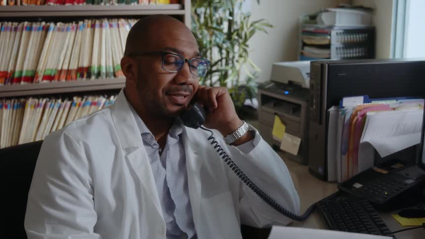 A man wearing a white lab coat is sitting at a desk as he holds a telephone receiver and reads a document