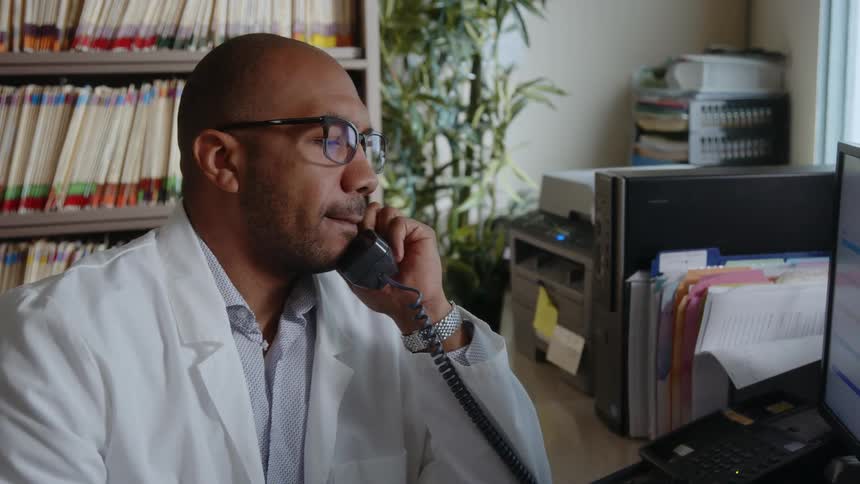 Doctor sitting in office near computer and coded files makes a phone call