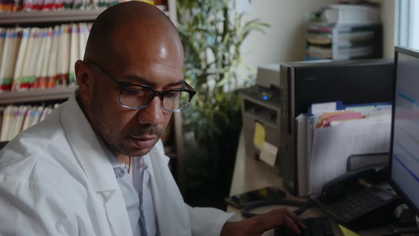 A man wearing a white lab coat is sitting at a desk typing on a computer keyboard
