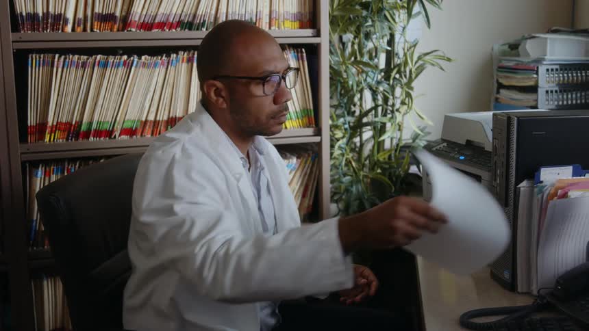 A man wearing a white lab coat is sitting at a desk in front of a computer holding a document as he rolls his chair over to the copy machine and places the document inside