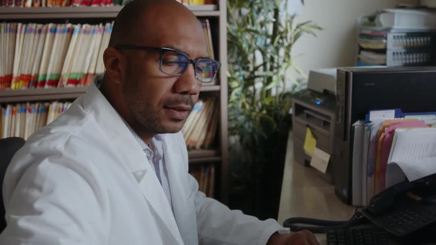 A doctor sitting at a desk has a drink from a thermal mug then puts it down and moves to work on a computer