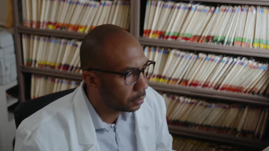 A black office chair a male doctor sits in is his office with files on the wall behind him