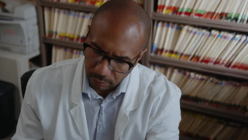 A man wearing a white lab coat is sitting at a desk moving his head as he works