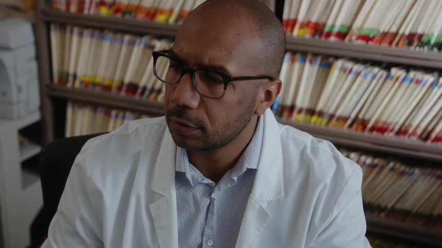 Male doctor sits in room near shelves full of color coded files and works