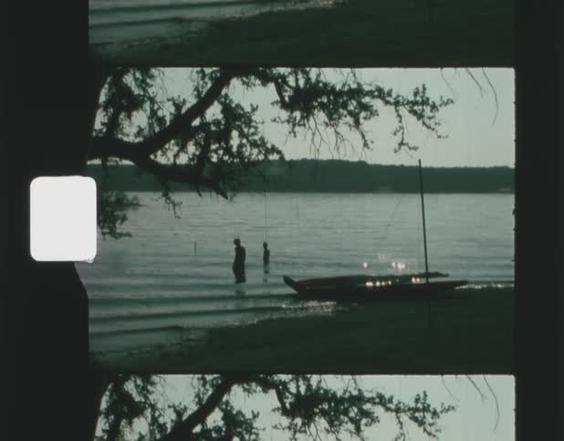 An adult and child are wading in a river next to a sailboat on the shore