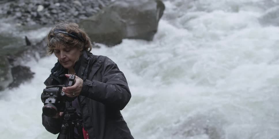 A woman adjusts her camera in front of a river