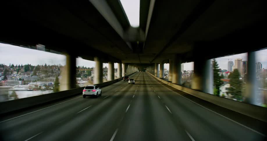 Traffic moves on a freeway in and out of town and through an underpass toward with buildings and skyscrapers in the distance under a cloudy sky