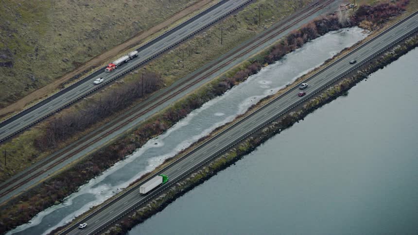 A very long and empty road with cars driving on both sides with water on one side and grass surrounding the peaceful scene