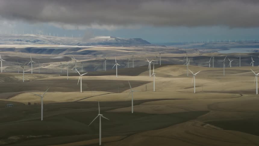 Aerial shot of Wind Turbines rotating above storm clouds in dappled light with a large river in the background