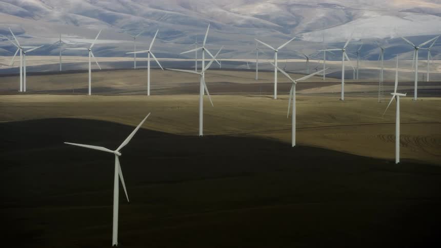 A large wind farm with white windmills on the landscape with mountains in the distance behind in this high altitude aerial shot