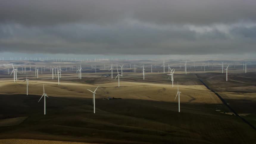 An aerial of windmills and wind turbines spinning on a large plateau of dry land wheat with moody storm clouds floating in the sky above