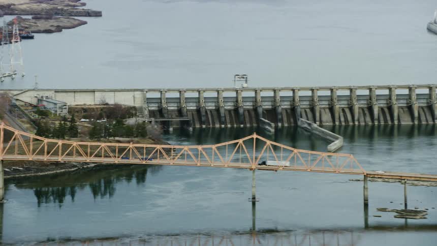 An aerial of a waterway and dam near a bridge going over way with traffic traveling on it