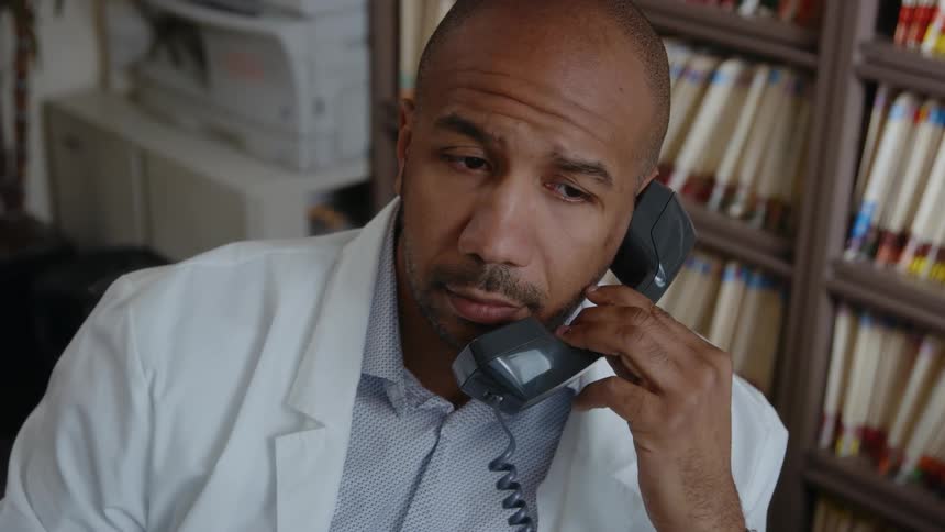 A close up of a male doctors face while he talks on the phone sitting next to a wall of files