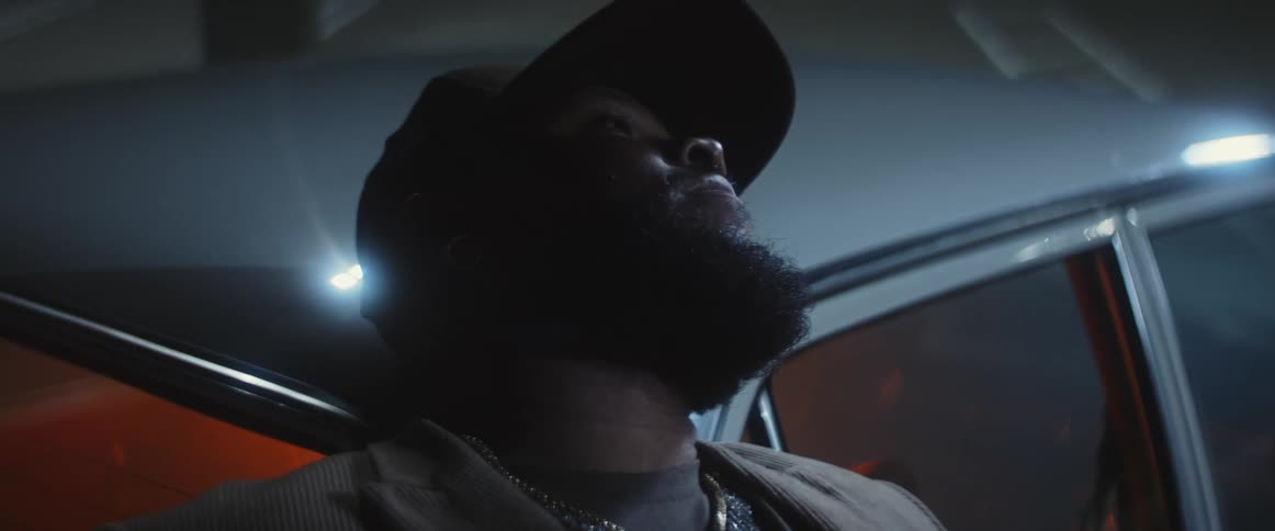 A man wearing a baseball cap is leaning against a car and looking up