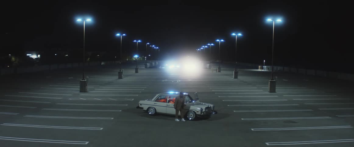 A man waits outside of his vehicle that is parked in an empty parking lot while other vehicles are approaching at night