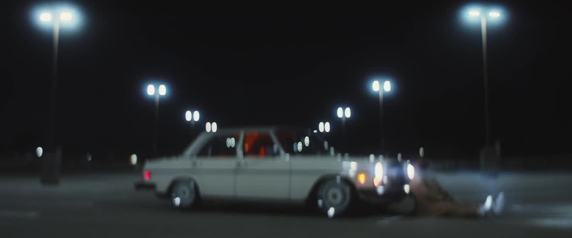 A man sits against the front of his car in an empty parking lot