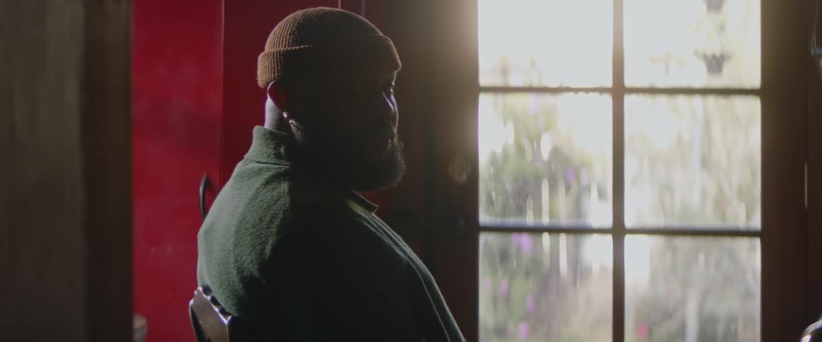 A black man is sitting in a chair looking contemplative and sad and then looks to his side and towards the camera