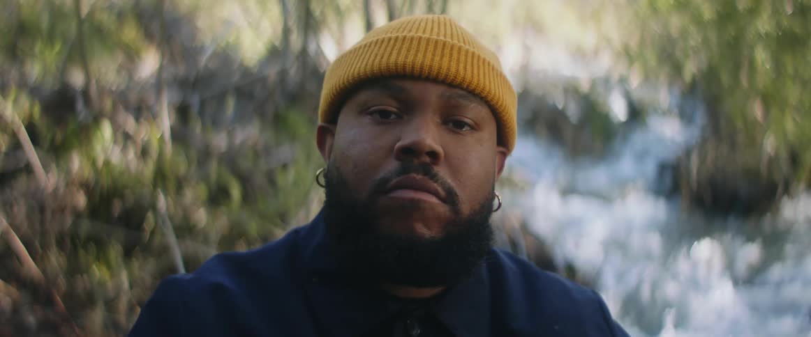 Man with yellow beanie earrings and beard stares at camera near the stream