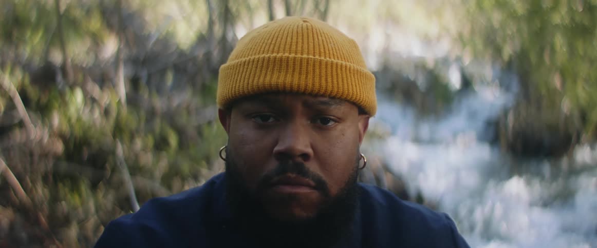 A man in a beanie poses for the camera in front of a creek